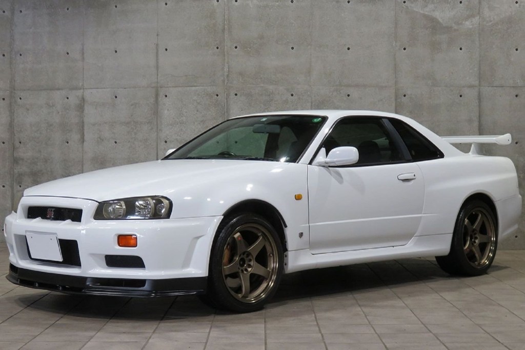 A white 1999 R34 Nissan Skyline GT-R by a gray stone wall
