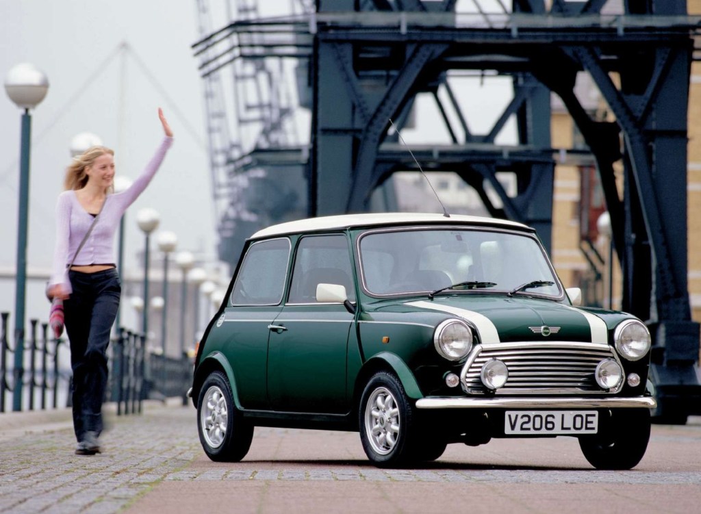 A woman walks next to a green-with-white-stripes 2000 Rover Mini Cooper by a European bridge
