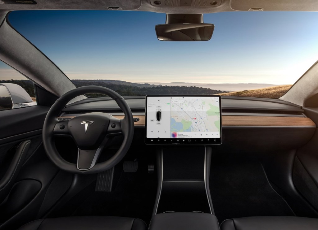 The black front seats and wood-trimmed dashboard of a 2018 Tesla Model 3 showing the large screen that is used to control the Tesla Autopilot software
