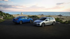 A blue 2020 Subaru Impreza sedan and a silver 2020 Subaru Impreza hatchback in a parking lot overlooking a beach