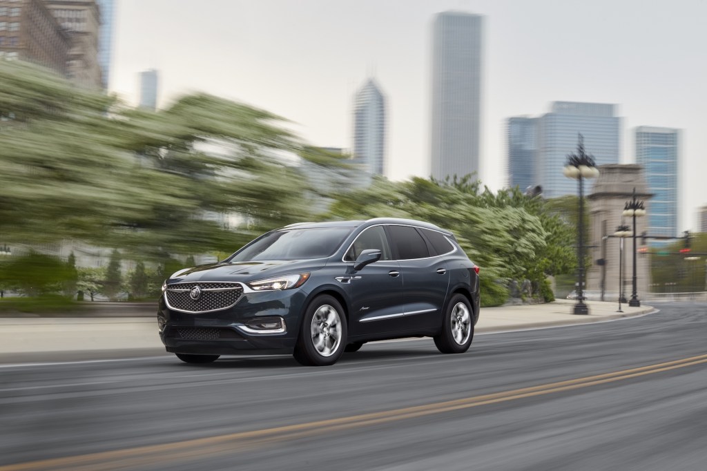 A dark-blue 2021 Buick Enclave luxury midsize crossover SUV traveling on a city street lined with trees and lamp posts
