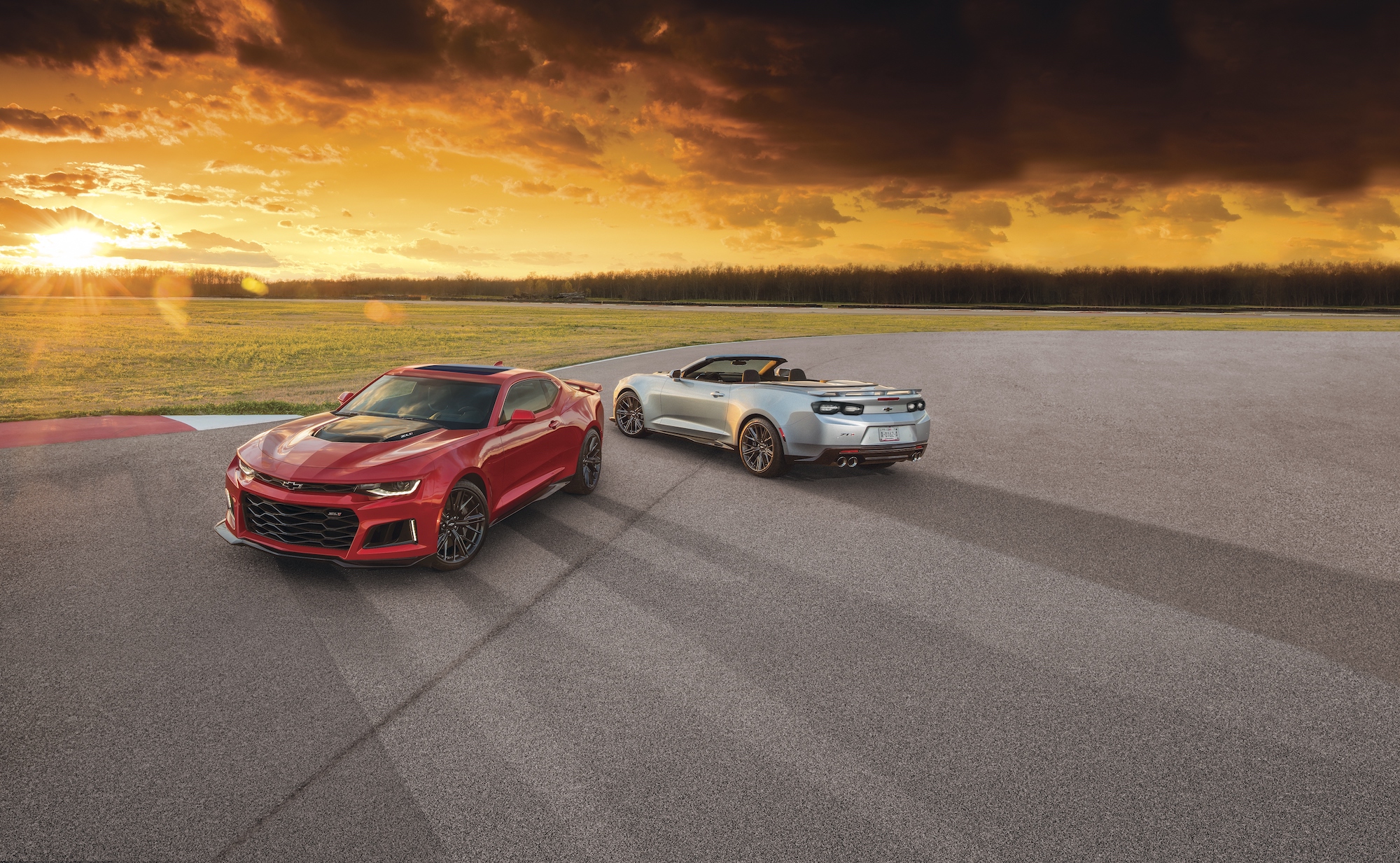 A red 2021 Chevy Camaro ZL1 coupe and a silver 2021 Chevy Camaro ZL1 convertible parked on a large expanse of asphalt overlooking grass, trees, and the sun on the horizon