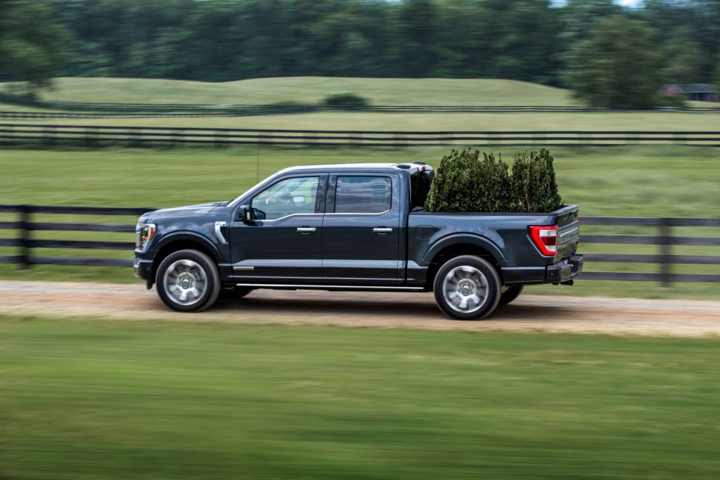 A dark-blue 2021 Ford F-150 PowerBoost Hybrid drives down a field road carrying some trees