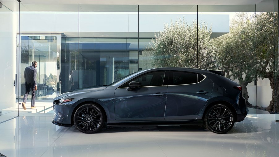 A bluish-gray 2021 Mazda3 2.5 Turbo hatchback sits in a white glossy showroom as a man in a business suit walks away