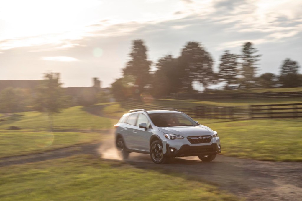 A pearl-white 2021 Subaru Crosstrek subcompact SUV traveling on a dusty country road along rolling hills and brown wooden fences