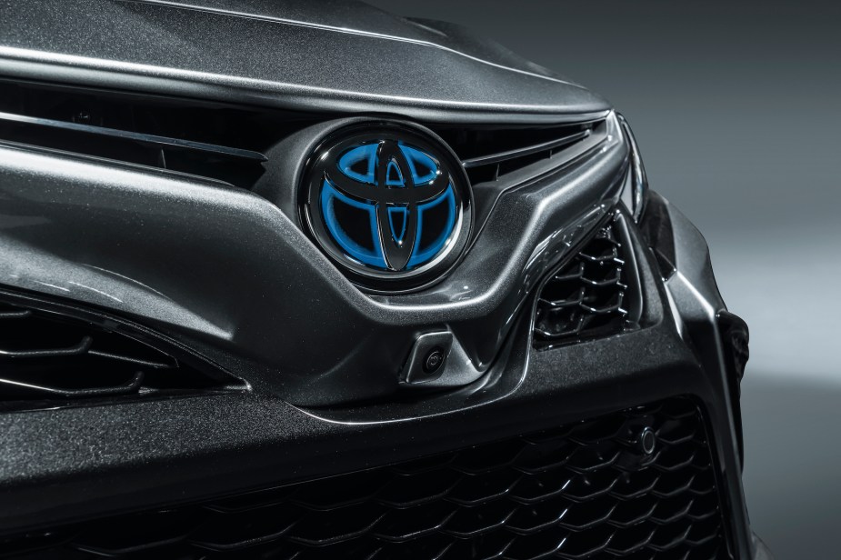 A dark-gray metallic 2021 Toyota Camry Hybrid sedan's honeycomb grille bears a silver and blue Toyota logo