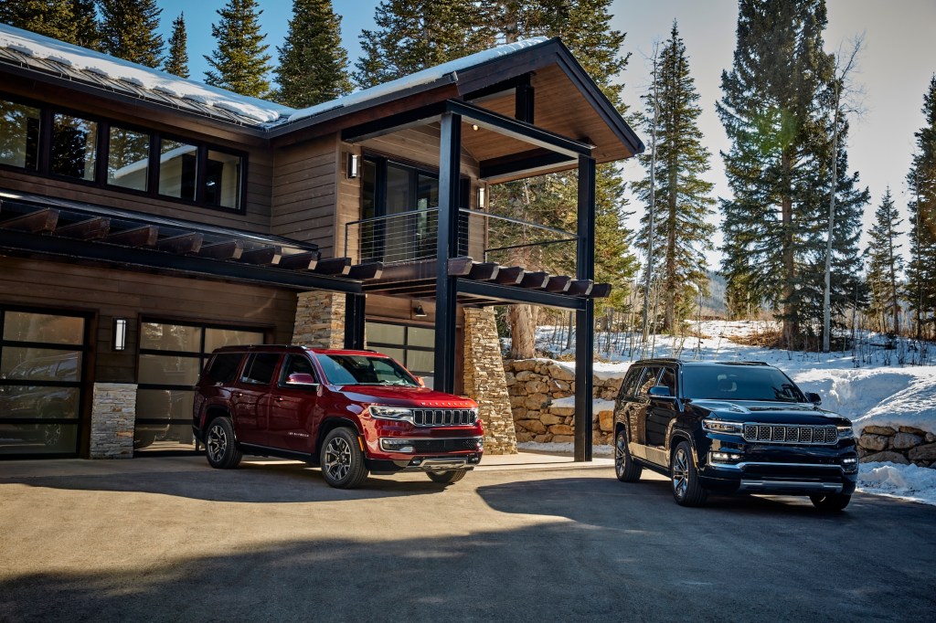 A red 2022 Jeep Wagoneer and a dark-colored 2022 Jeep Grand Wagoneer parked in front of a modern mountain home surrounded by snow and pine trees
