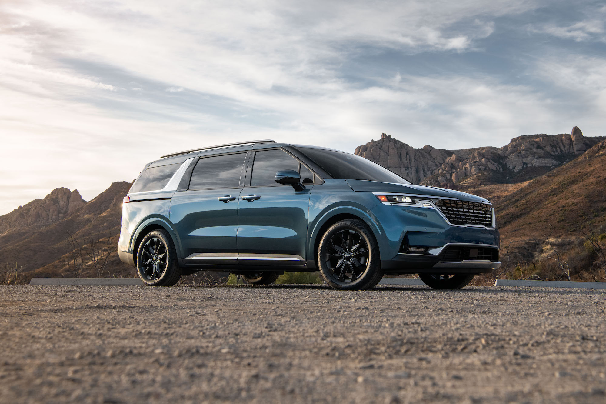A blue 2022 Kia Carnival minivan parked on gravel in front of mountains on a partly cloudy day