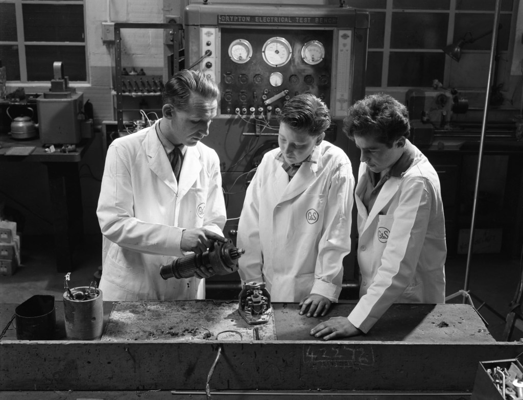 A mechanic trains two apprentices on a starter motor in a garage in 1961