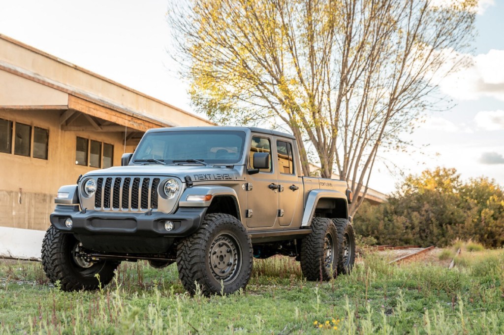 A matte-gray Next Level Jeep Gladiator 6x6 parked by a yellow-stone building's lawn