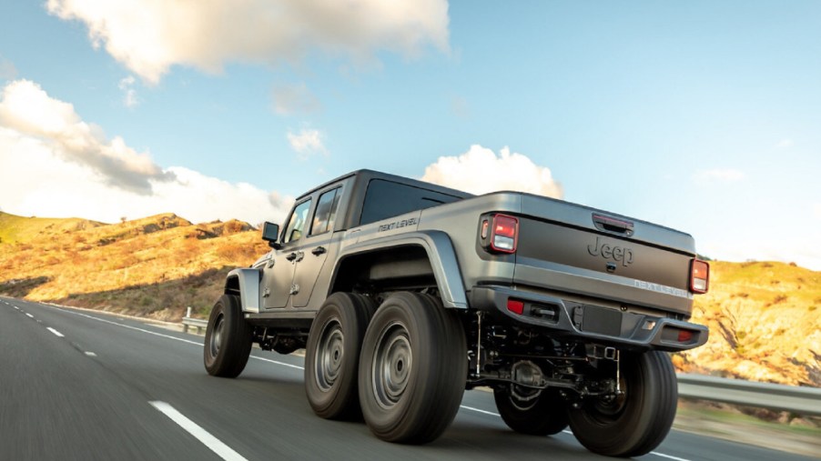 A low-angle rear 3/4 view of a matte-gray Next Level Jeep Gladiator 6x6 driving on a hilly road