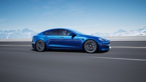 A royal-blue metallic Tesla Model S traveling on a multi-lane highway with snow-covered mountains and a blue sky in the background