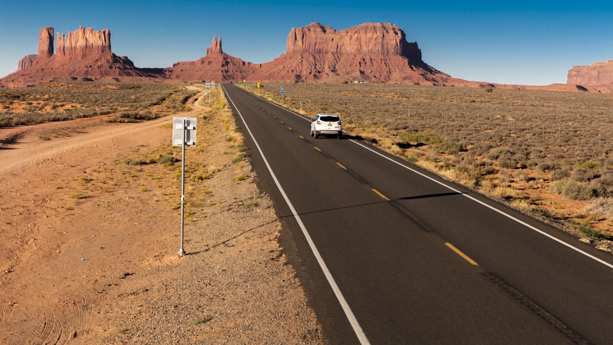 A white SUV on a road trip drives toward Monument Valley on the Utah-Arizona border in the American Southwest