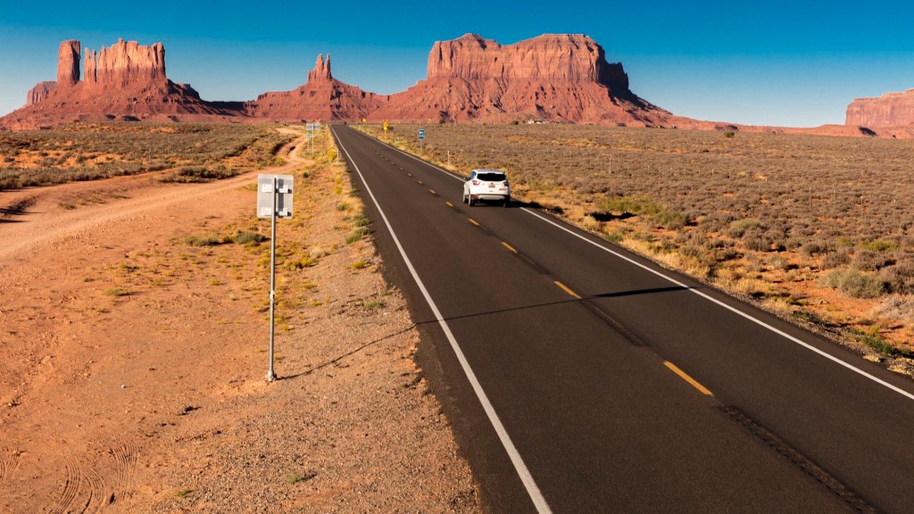 A white SUV on a road trip drives toward Monument Valley on the Utah-Arizona border in the American Southwest