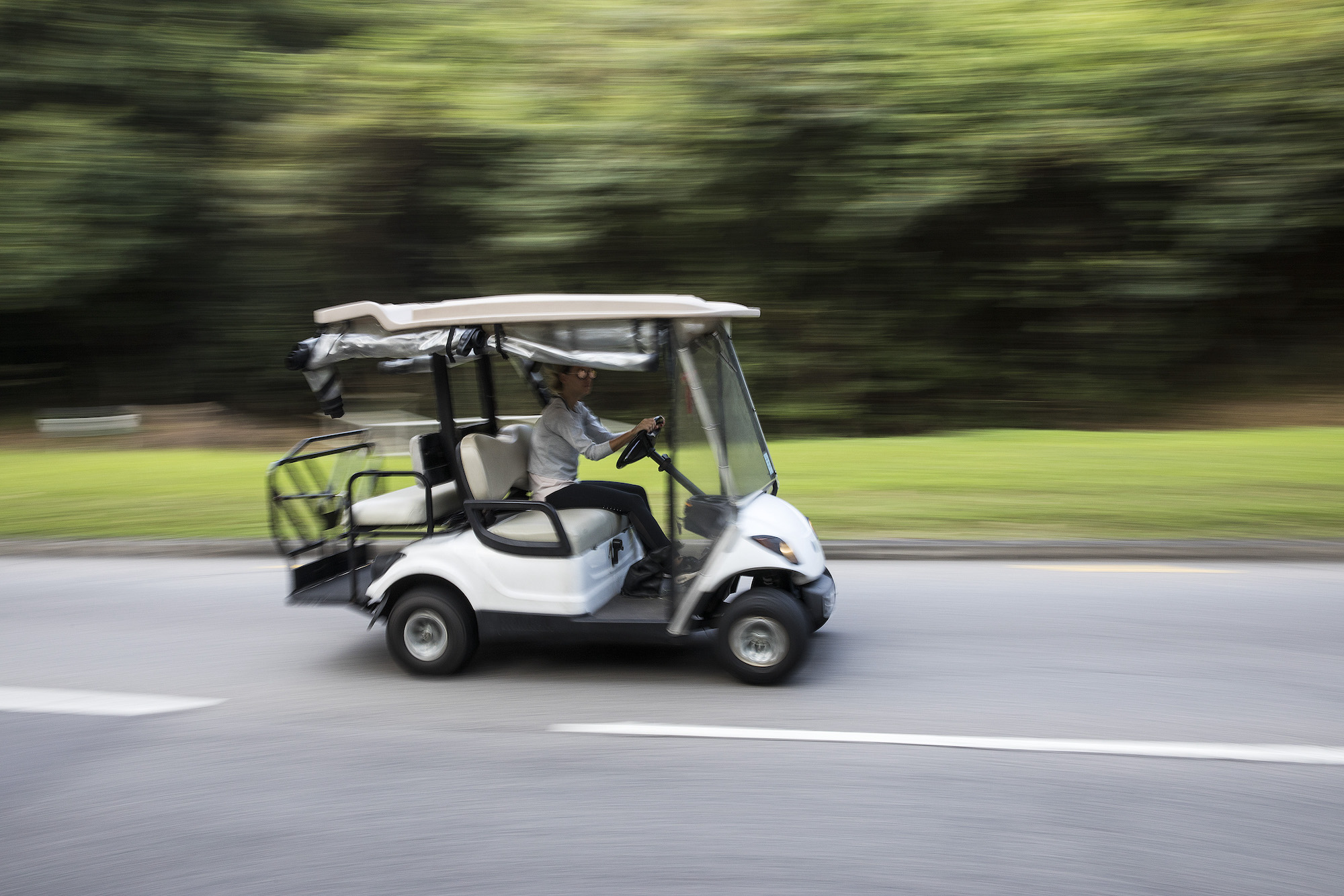 A white golf cart travels on a residential road along green grass and lush trees