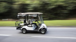 A white golf cart travels on a residential road along green grass and lush trees