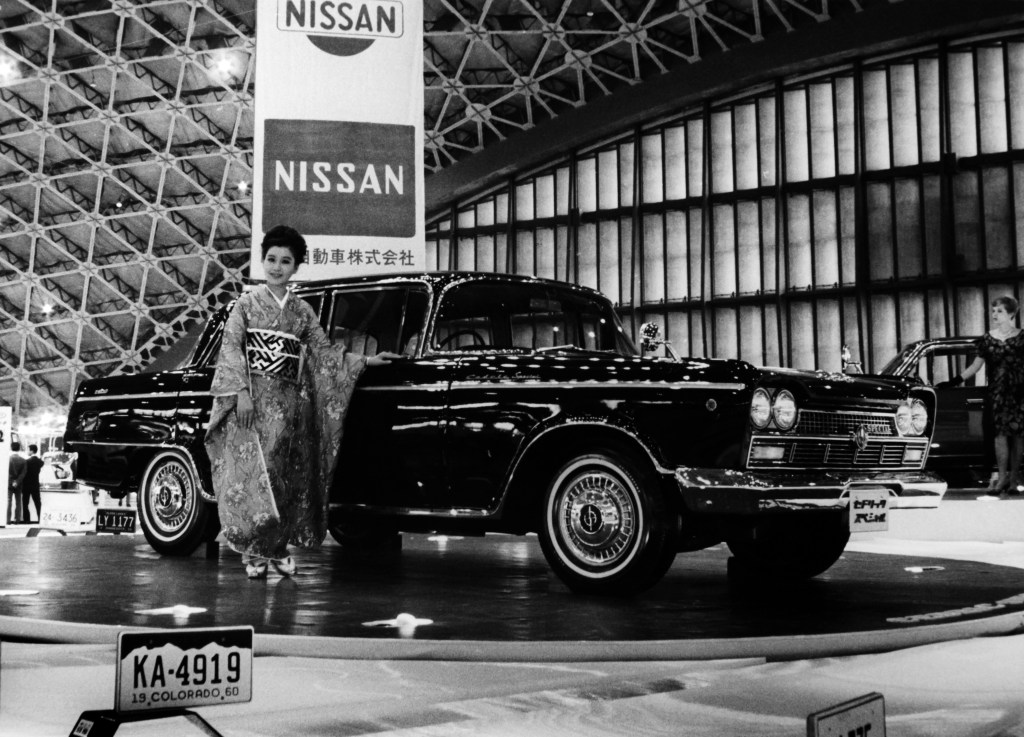 A black and white archival photo of a dark Nissan Cedric sedan being displayed by a woman in a kimono