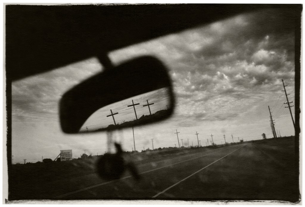View of three crosses on the side of a highway, as seen through the rearview mirror of a car.