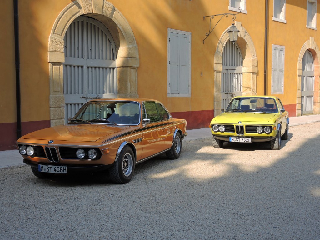 An orange and a yellow 1971 BMW 3.0 CSL by an orange-colored building