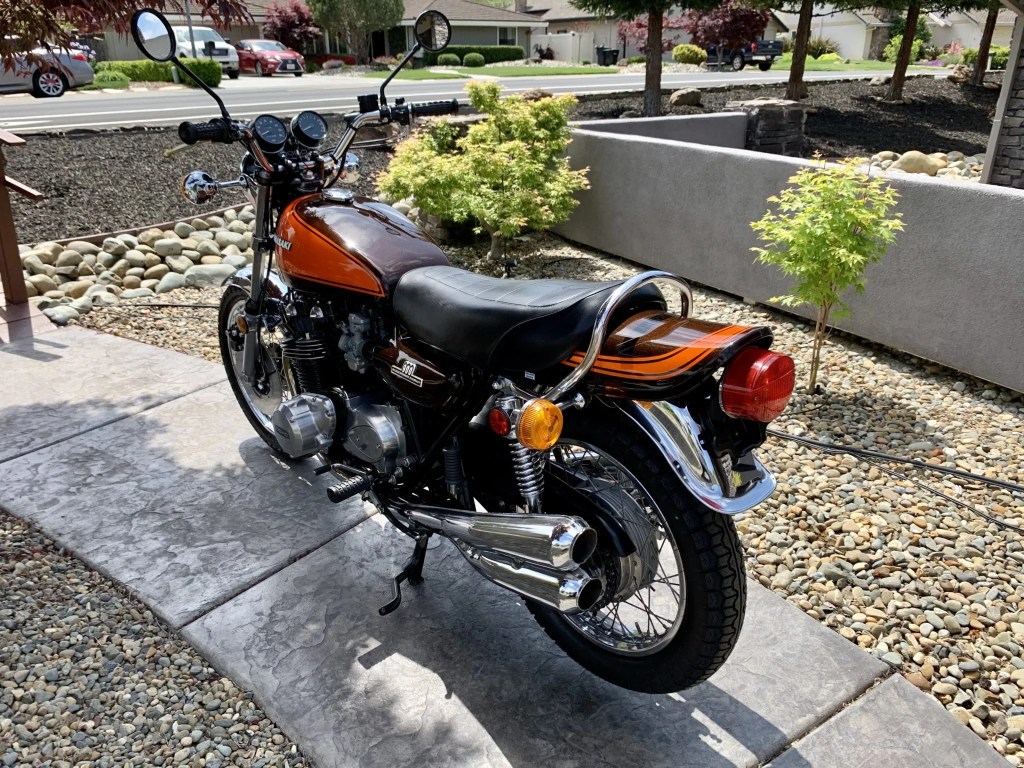 The rear 3/4 view of an orange-and-brown 1973 Kawasaki Z1 900 on a walkway in a rocky lawn