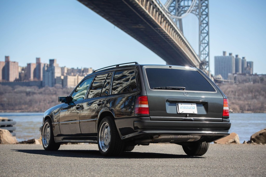 The rear 3/4 view of a black 1993 W124 Mercedes-Benz 320TE Wagon with the AMG kit by a NYC bridge