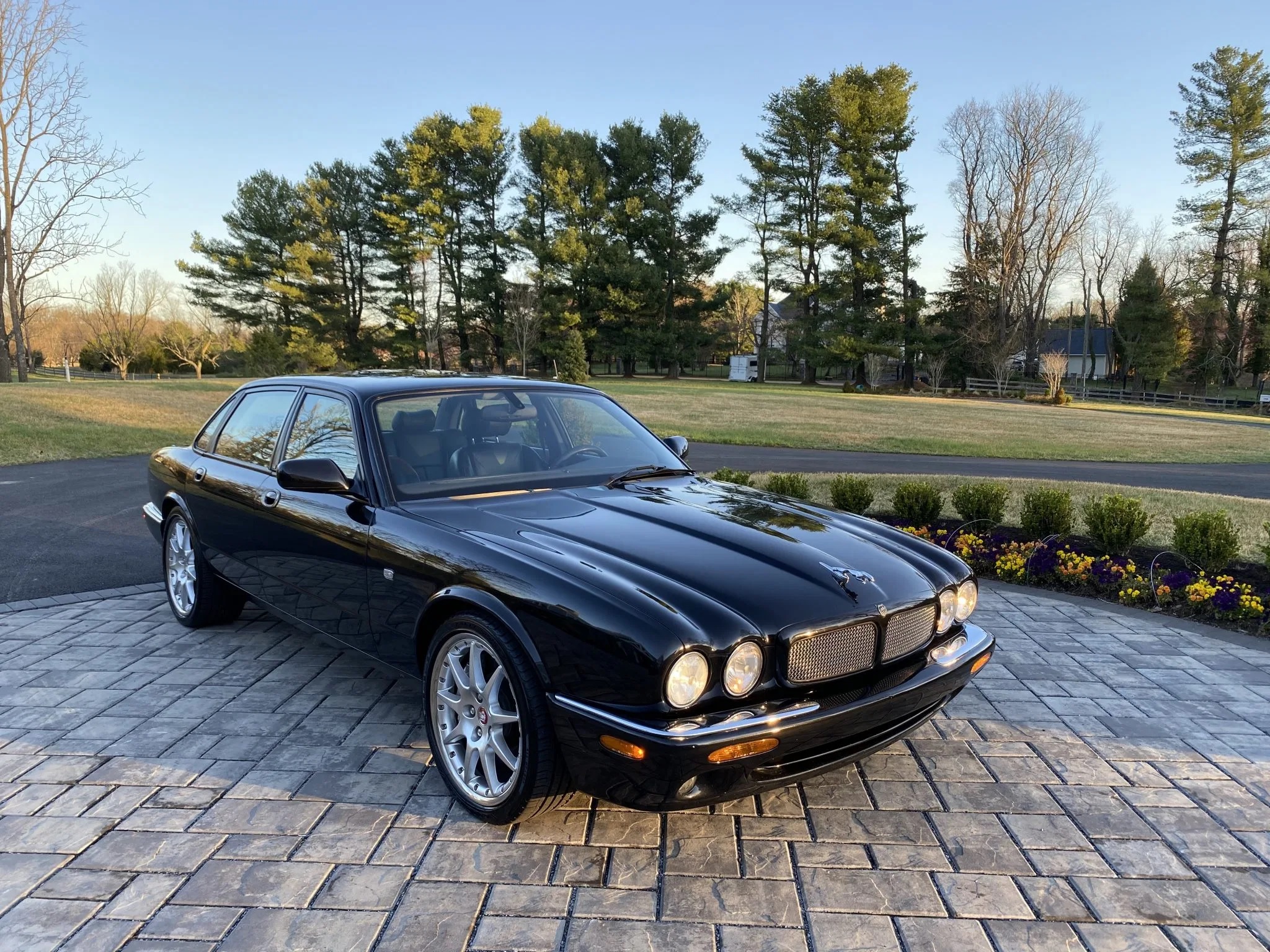 A black 2002 Jaguar XJR 100 on a stone driveway in front of a tree-lined lawn
