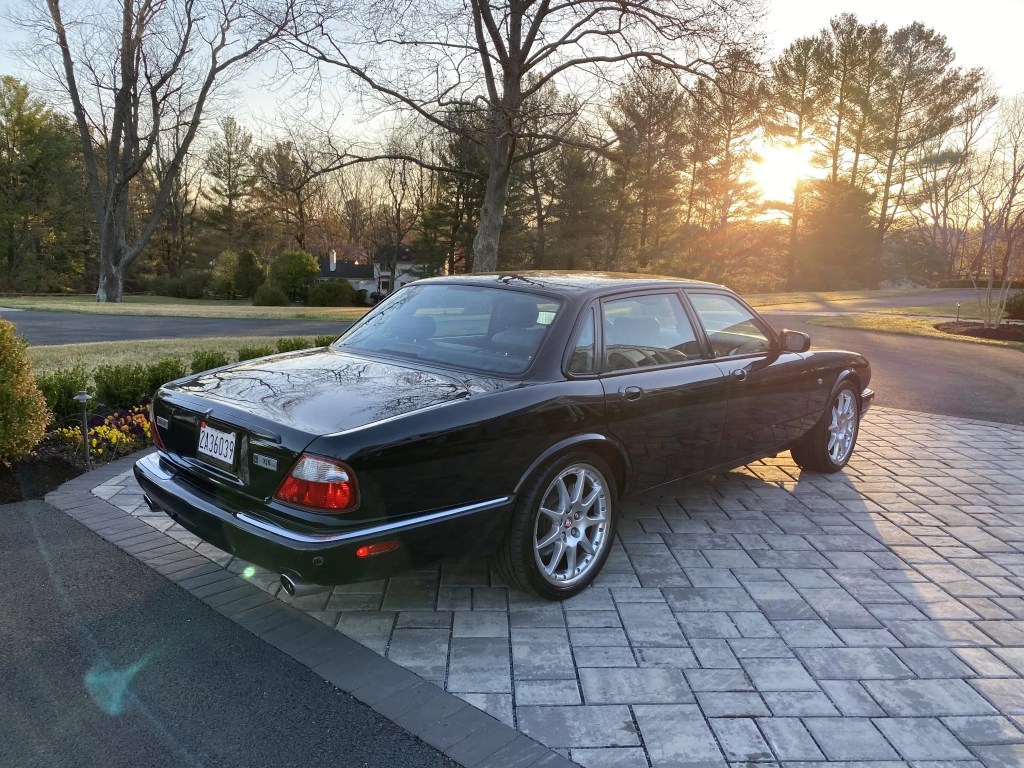 The rear 3/4 view of a black 2002 Jaguar XJR 100 on a stone driveway in front of a tree-lined lawn