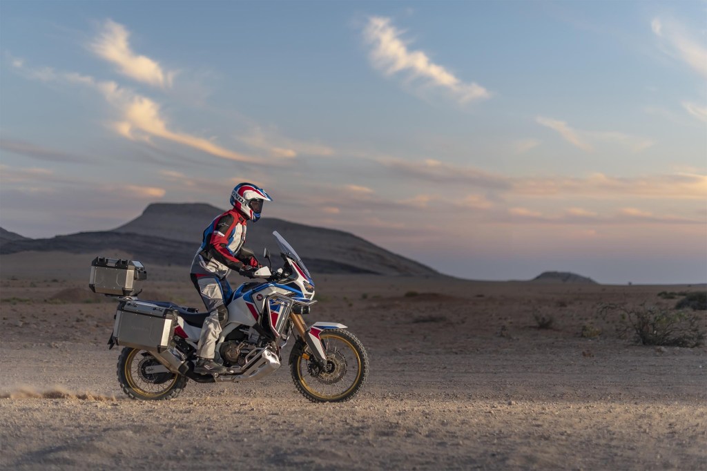 The side view of a white-red-and-blue-clad rider taking a white-red-and-blue 2020 Honda Africa Twin Adventure Sports ES DCT through a desert