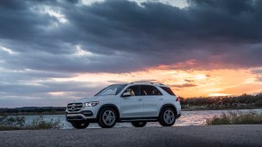 A white 2020 Mercedes-Benz GLE midsize luxury SUV parked overlooking a body of water with dark clouds overhead and the sun on the horizon