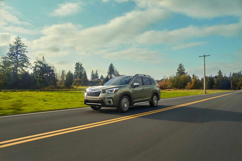 A sage-green 2020 Subaru Forester traveling on a two-lane highway past green grass and pine trees on a partly cloudy day, one of the safest compact SUVs recommended by Consumer Reports.