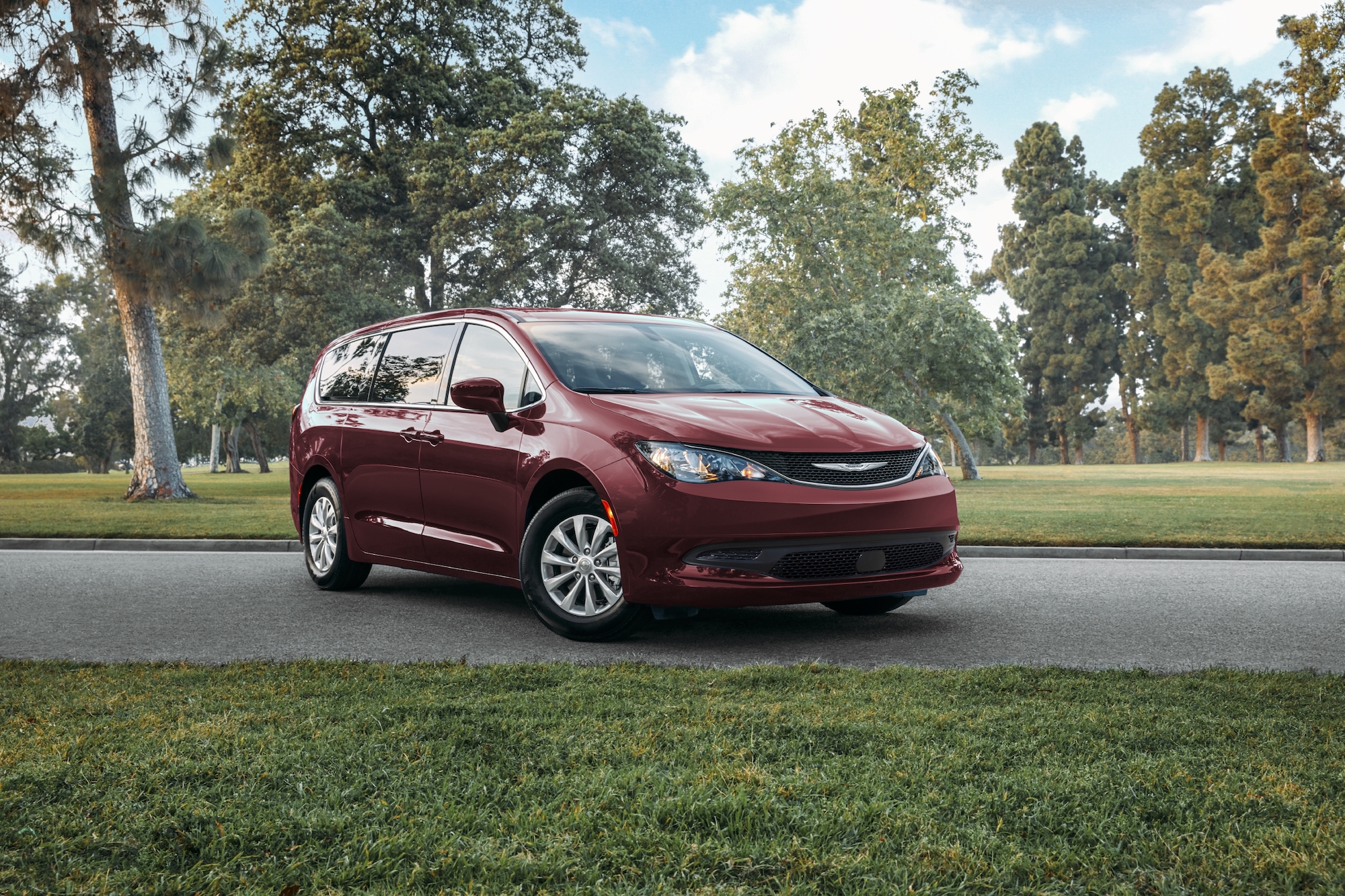 A dark-red 2021 Chrysler Voyager minivan parked on a suburban street surrounded by green grass and trees on a mostly sunny day