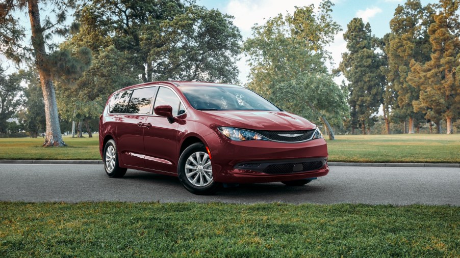 A dark-red 2021 Chrysler Voyager minivan parked on a suburban street surrounded by green grass and trees on a mostly sunny day