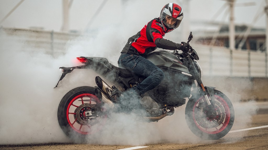 A red-clad rider on a gray 2021 Ducati Monster does a burnout on a racetrack