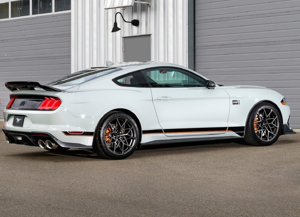 The side 3/4 view of a gray-black-and-orange 2021 Ford Mustang Mach 1 with the Handling Package by a racetrack garage
