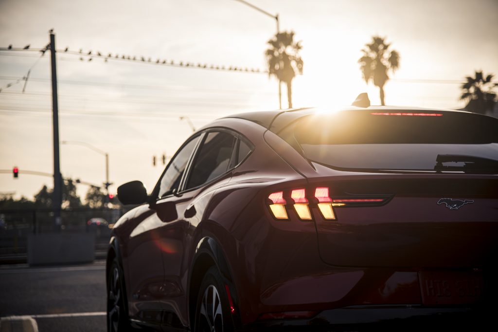 A rear view of a red 2021 Ford Mustang Mach-E electric SUV stopped on a street lined with palm trees as the sun hangs over the horizon