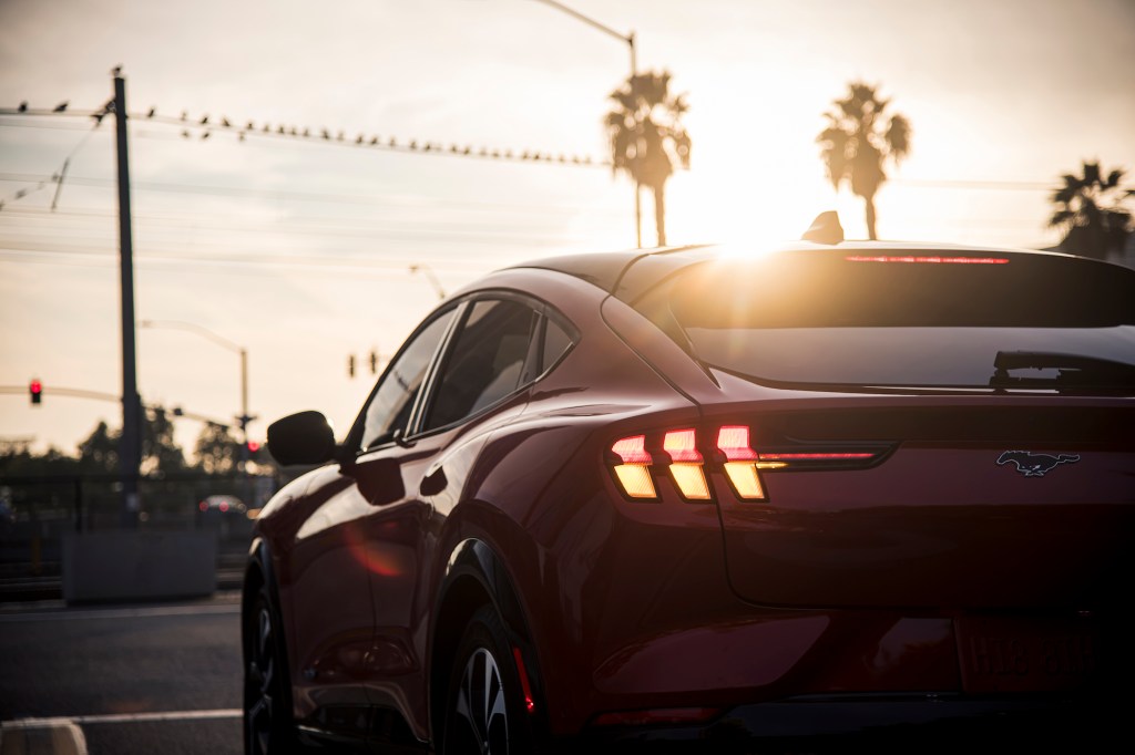 A rear view of a red 2021 Ford Mustang Mach-E electric SUV stopped on a street lined with palm trees as the sun hangs over the horizon
