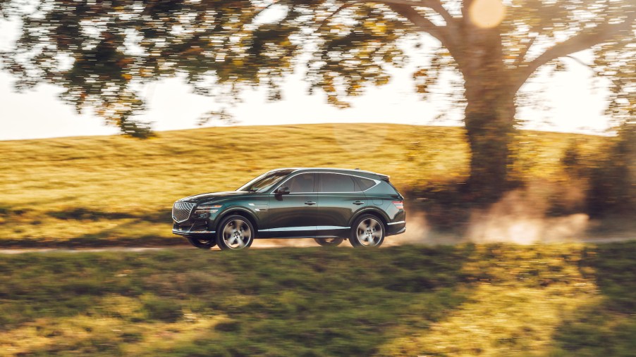 A dark-green 2021 Genesis GV80 midsize SUV traveling on a dusty road through rolling green hills in Hudson Valley, New York