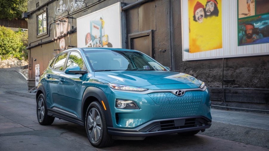A turquoise 2021 Hyundai Kona Electric crossover SUV parked on a street outside a black building with Cheech & Chong artwork on it