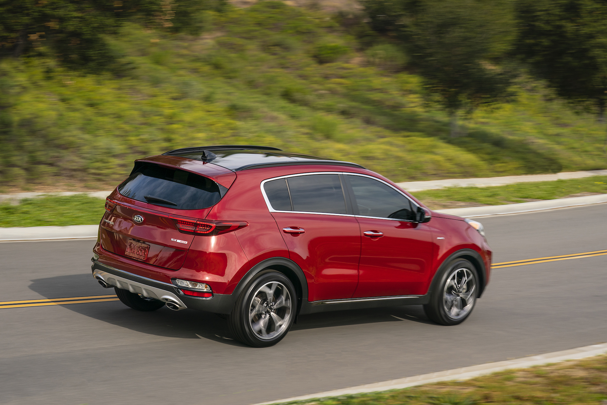 A red 2021 Kia Sportage compact SUV travels on a two-lane highway on a grassy, tree-dotted hill