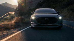 A dark-gray metallic 2021 Mazda3 2.5 Turbo compact car traveling on a two-lane highway next to a mountain