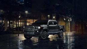 A black 2021 Toyota Tacoma Nighshade midsize pickup truck parked on wet pavement outside a glass building at night