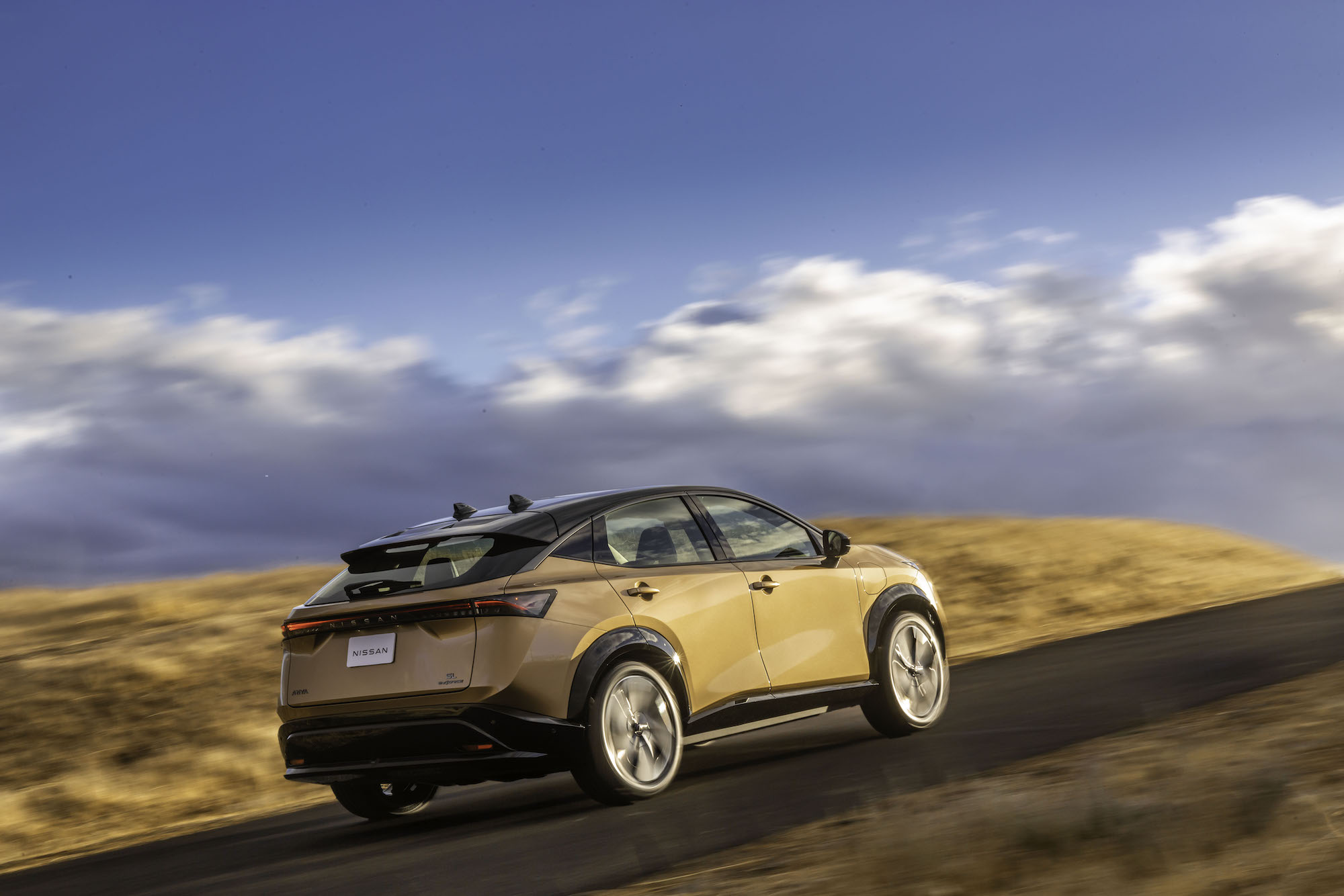 A gold 2022 Nissa Ariya electric compact SUV traveling uphill on a partly sunny day