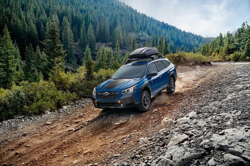 A blue 2022 Subaru Outback Wilderness driving down a dirt road near the forest