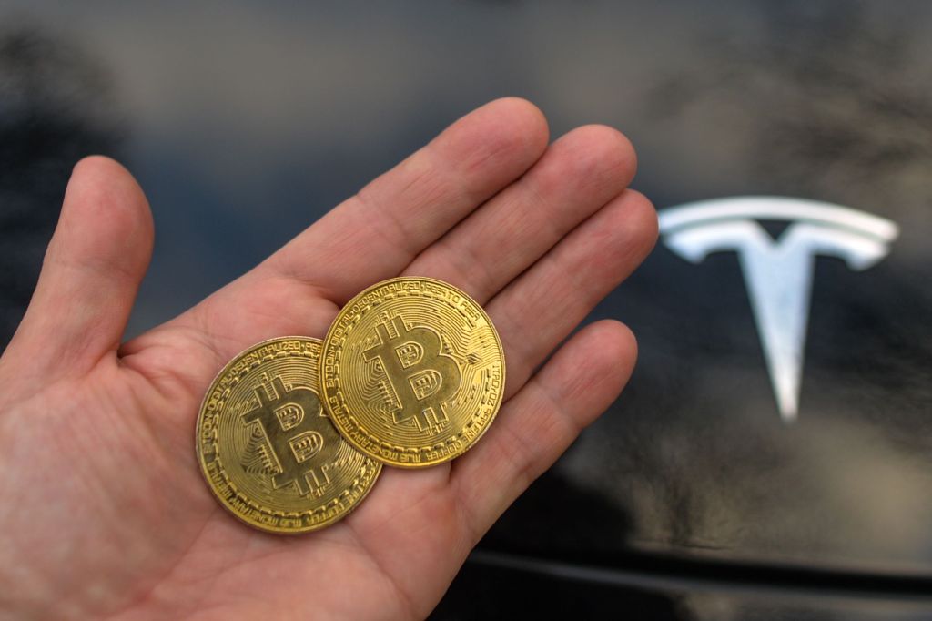 A person holds two commemorative Bitcoin coins next to a black Tesla EV