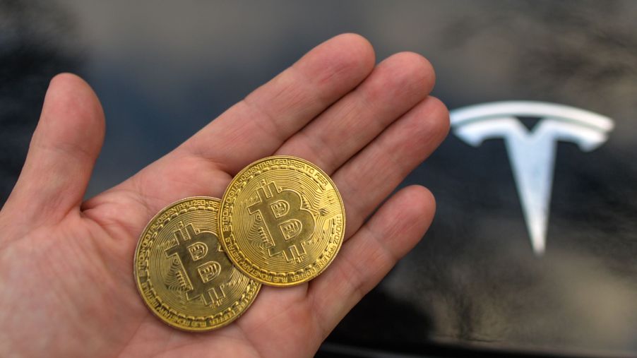 A person holds two commemorative Bitcoin coins next to a black Tesla EV