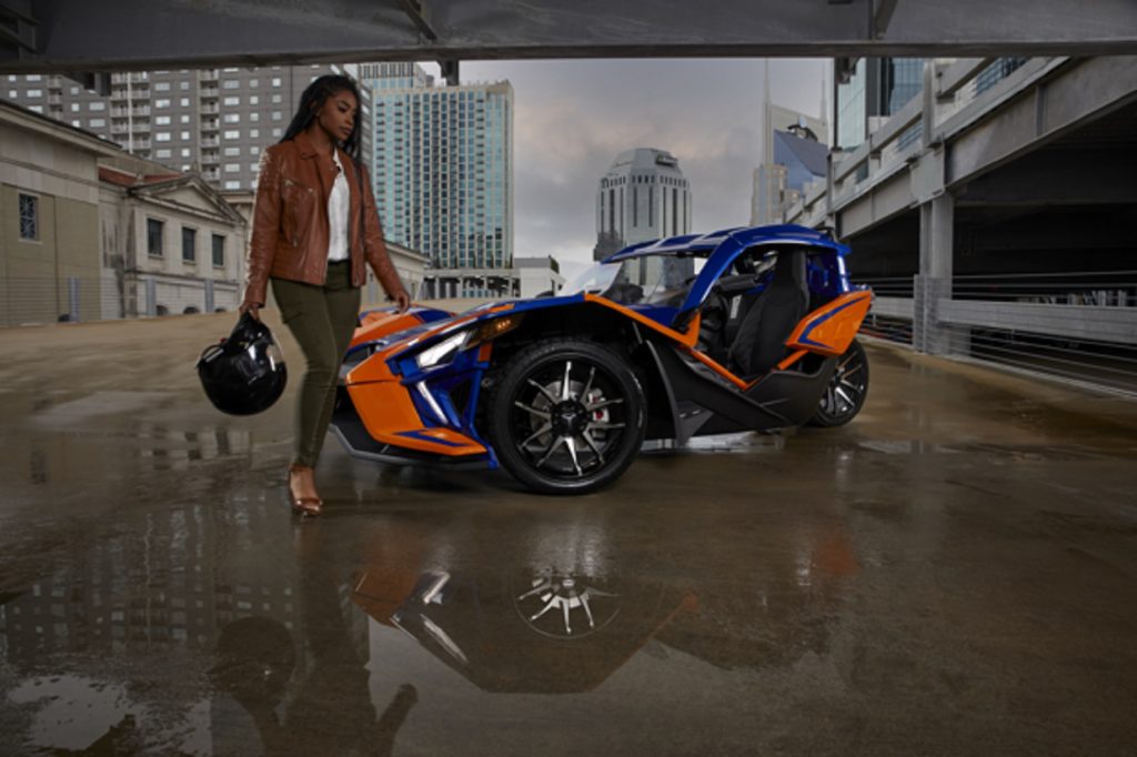 A woman in a brown-leather jacket with a black helmet in hand walks up to a blue-and-orange 2021 Polaris Slingshot underneath a rainy city bridge