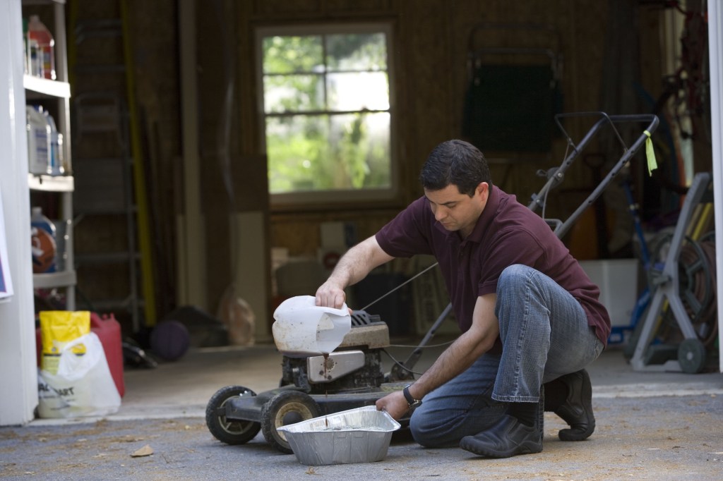 Man in the process of changing his lawnmower's oil outside a garage, using a disposable aluminum catch pan. 