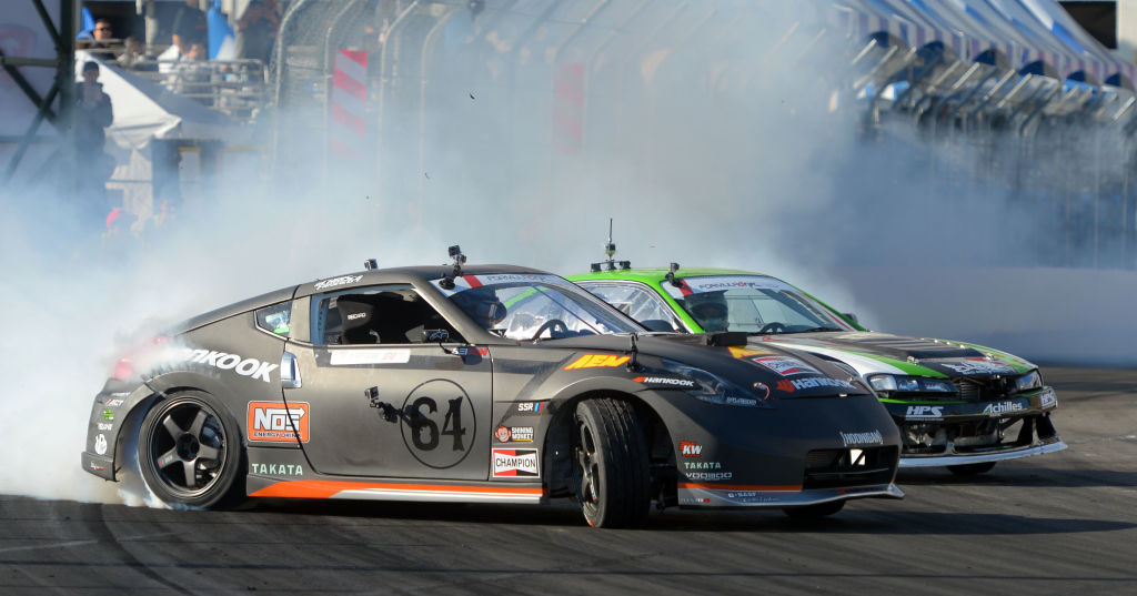 Two Formula Drift cars slide out of a corner spewing smoke