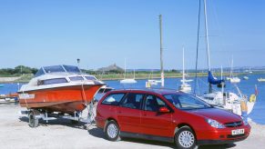 A red 2002 Citroen C5 hdi crossover towing a boat by a harbor