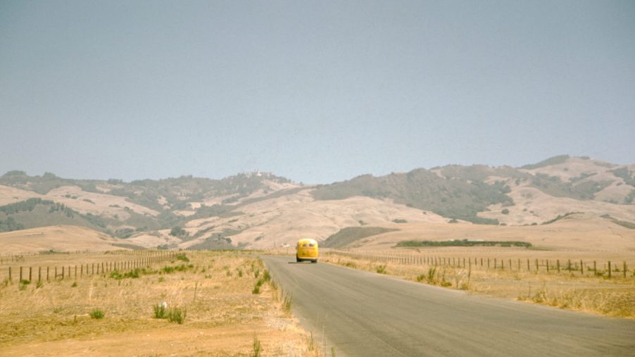 A yellow Volkswagen bus travels along a country road that cuts through a prairie covered in dry grass, low rolling hills can be seen in the background on a summer road trip